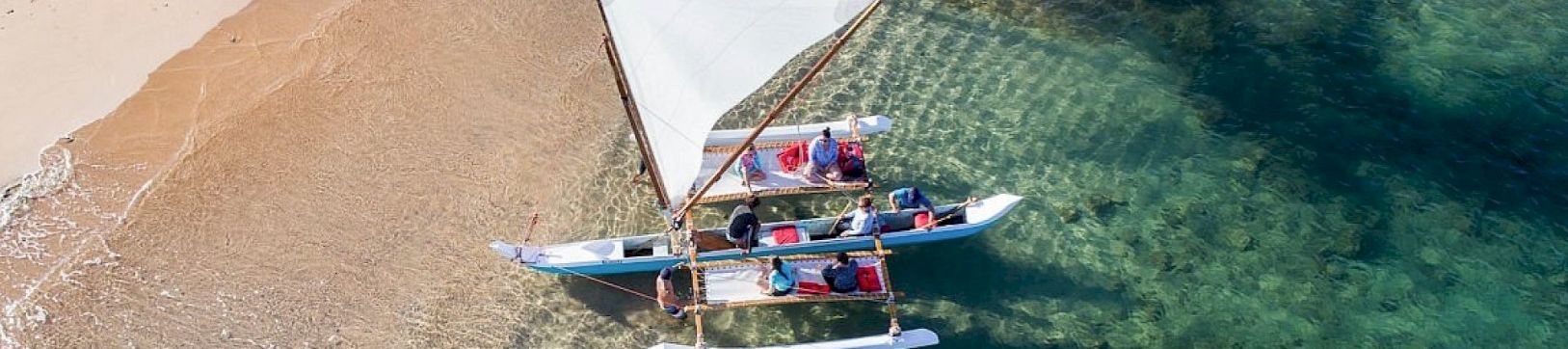 An overhead view of a small sailboat gliding on clear turquoise water near a sandy shore, with crew handling sails.