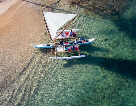An overhead view of a small sailboat gliding on clear turquoise water near a sandy shore, with crew handling sails.