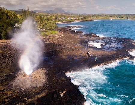 A coastal cliff with steam erupting from lava rocks as people stand near the waves under a cloudy sky.