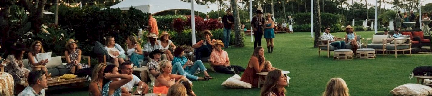 An outdoor tropical garden gathering with guests lounging on the grass under white tents, surrounded by tall palm trees.