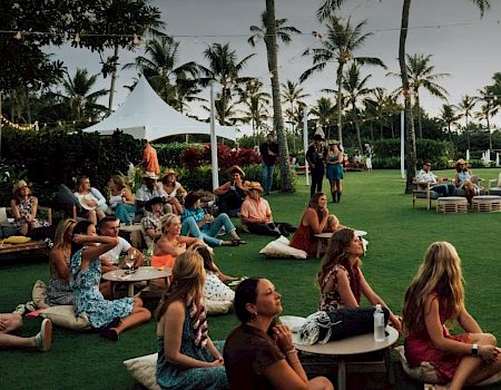 An outdoor tropical garden gathering with guests lounging on the grass under white tents, surrounded by tall palm trees.
