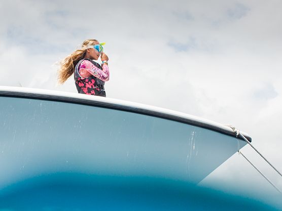 A person with long hair wearing a patterned hoodie leans from the open car window against a cloudy sky.