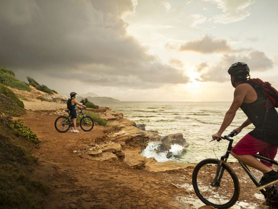 A couple of cyclists ride along a rocky seaside cliff as waves crash below and the sun breaks through clouds.