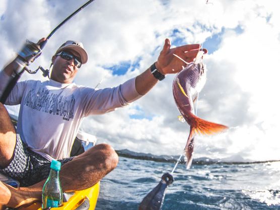 A fisherman on a boat reels in a red fish while ocean waves and blue sky fill the background.