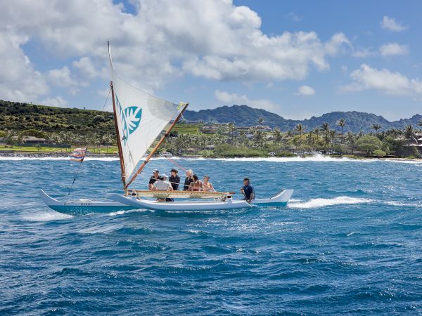 Sailboat with a sail and crew glides over sunny blue water near a tropical shoreline and mountains under cloudy skies.