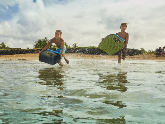Two kids splash in shallow ocean, surfing small boards and water-logging in the sunny beach surf.