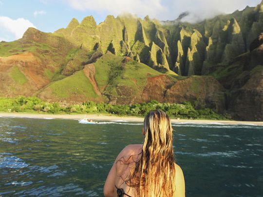 A woman with long hair sits by dark ocean water as a lush green mountain range rises under cloudy skies.