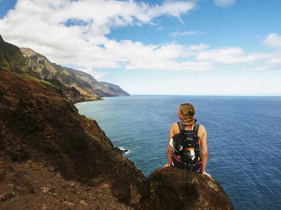 A hiker with a backpack sits on a cliff, overlooking the ocean and rugged mountains under a cloudy blue sky.