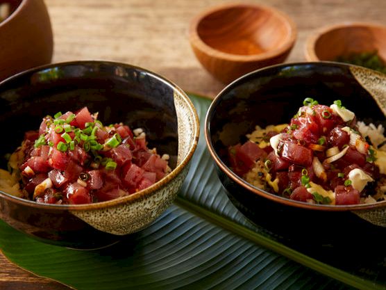 Two bowls of diced tuna poke/tartare over rice with scallions, sesame, and seaweed, on a wooden table and banana leaf.