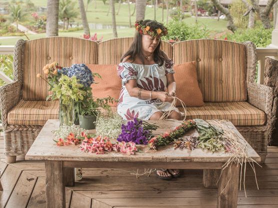 A woman wearing a flower crown sits on a porch swing with colorful flowers on a wooden table in front of her.