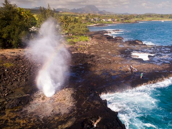 A volcanic steam plume rises over rocky black lava as ocean waves crash along a tropical coastline.