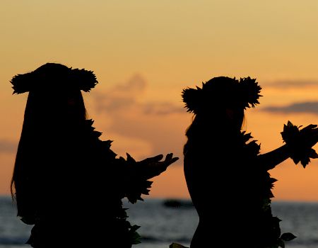 Silhouettes of two people wearing sunflower crowns on a beach at sunset, hands reaching out against the orange sky.
