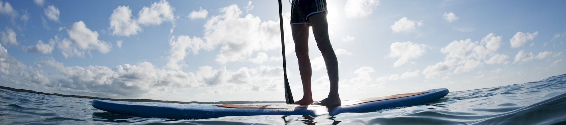 A person stands on a paddleboard, paddling across the ocean under a bright blue sky and scattered clouds.