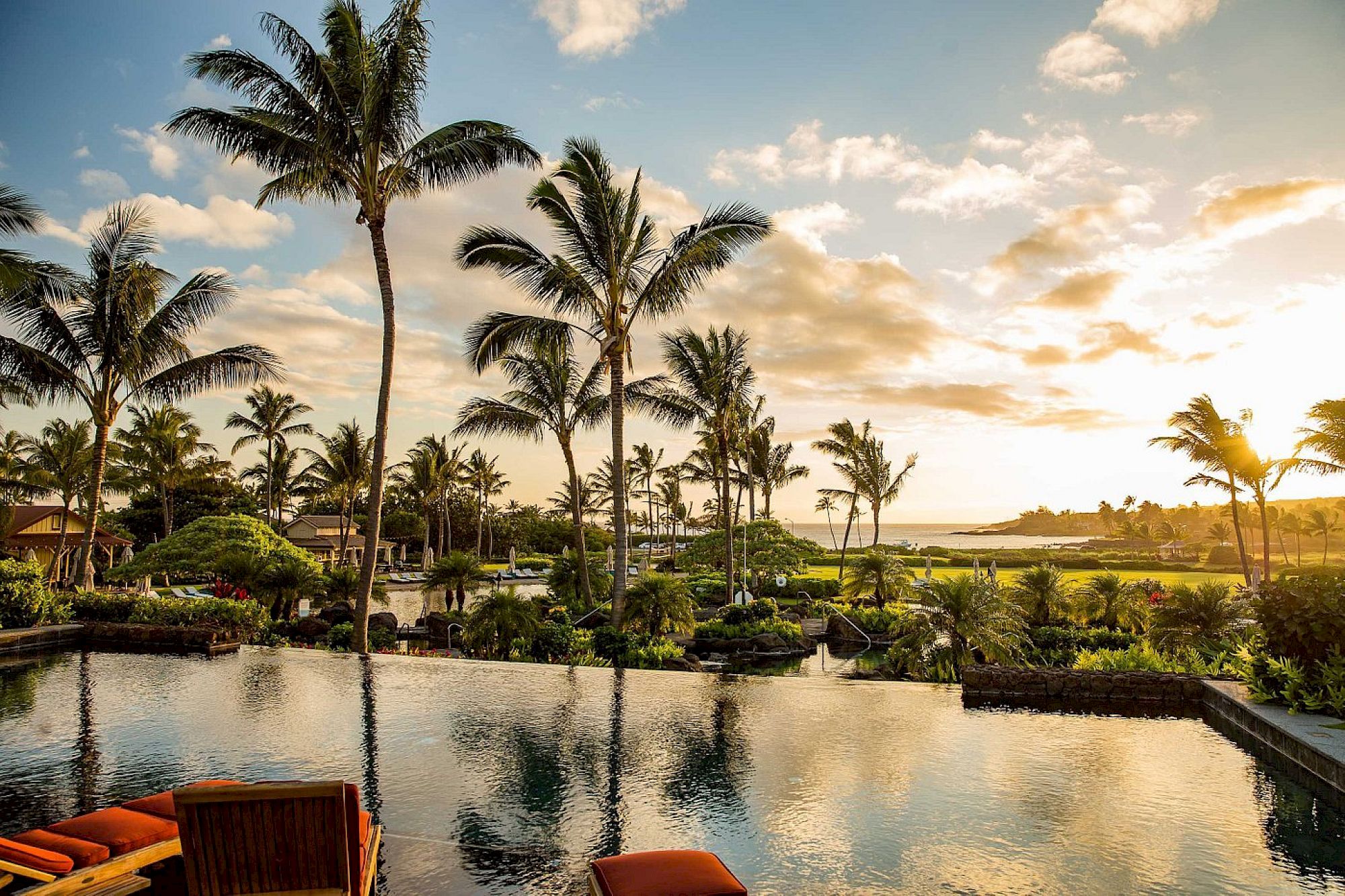 A tropical resort with tall palm trees, a calm pool/lagoon, and a warm sunset sky with clouds over the sea.
