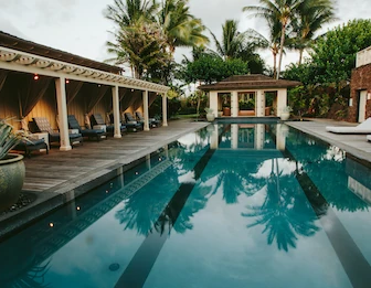 A tropical resort courtyard with a reflective swimming pool, lounge beds, palm trees, and a cozy wooden cabana area beside it.