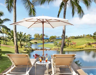 Two beige lounge chairs under a white umbrella on a tropical resort deck, overlooking a lagoon, palm trees, and the ocean.