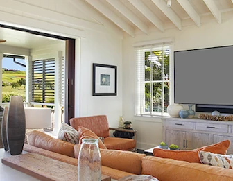 A bright living room with orange sectional seating, large windows, and a wall-mounted TV/screen under white beams.