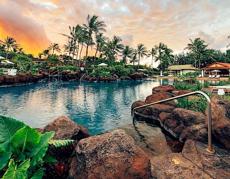 Tropical resort pool at sunset, bordered by rocks, palms, and cabanas with umbrellas.