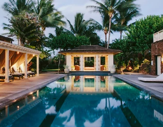 A tropical villa resort with a glowing swimming pool, lounge beds, and tall palm trees under a dusk sky.
