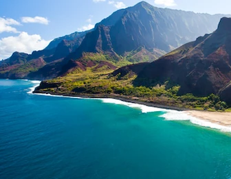 Aerial view of a turquoise coastline with a sandy beach, waves, and steep green mountains under a bright blue sky.