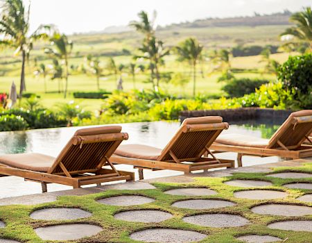 Sunlit tropical resort pool area with wooden lounge chairs, palm trees, and lush green hills in the background.