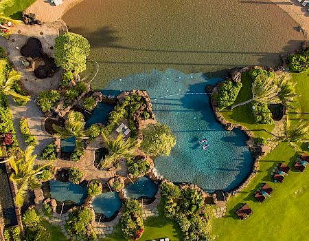 Aerial view of a tropical resort with multiple pools, sunbeds, palm trees, and stone landscaping.