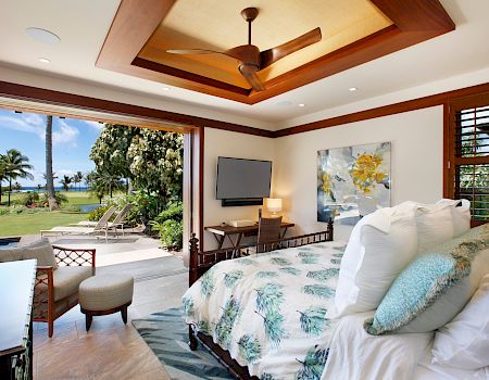 A luxury bedroom with a ceiling fan, big windows, TV, and a view of palm trees and ocean.