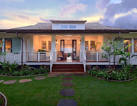 A white house with a welcoming front porch, steps, and landscaped garden under a colorful sunset sky.