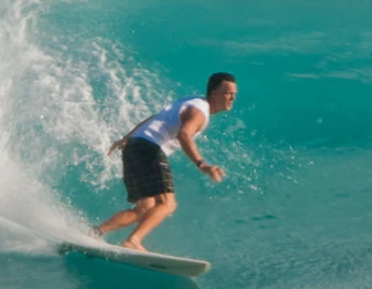 A surfer in a black swimsuit rides a turquoise wave on a surfboard, leaning forward as water splashes around the board.
