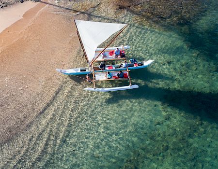 A small sailboat with several people floats near a sandy beach in clear turquoise water.