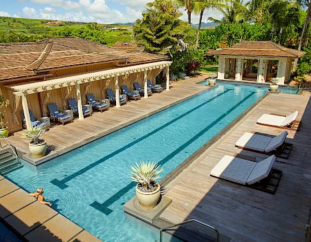 A sunny resort pool beside a landscaped patio, lounge chairs, and palm trees overlooking green hills and houses.