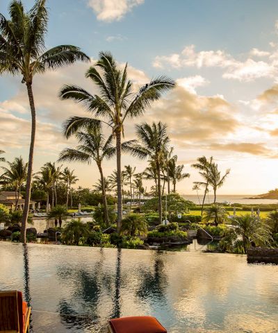 A tropical resort scene with tall palm trees, a calm pool reflecting sunset, and lush greenery under golden clouds.
