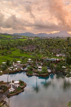 A tropical resort with palm trees, manicured lawns, and a lagoon-style pool at sunset, overlooking green hills and mountains.