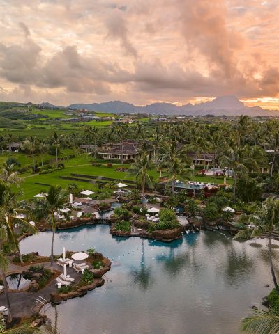 A tropical resort with palm trees, manicured lawns, and a lagoon-style pool at sunset, overlooking green hills and mountains.