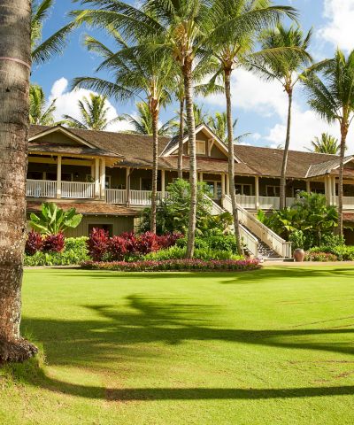 A tropical resort with a manicured green lawn, colorful garden beds, and palm trees surrounding a large multi-level building.