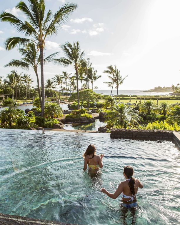 Palm-lined tropical resort with a clear pool, two children swimming, and the ocean under a sunny sky.