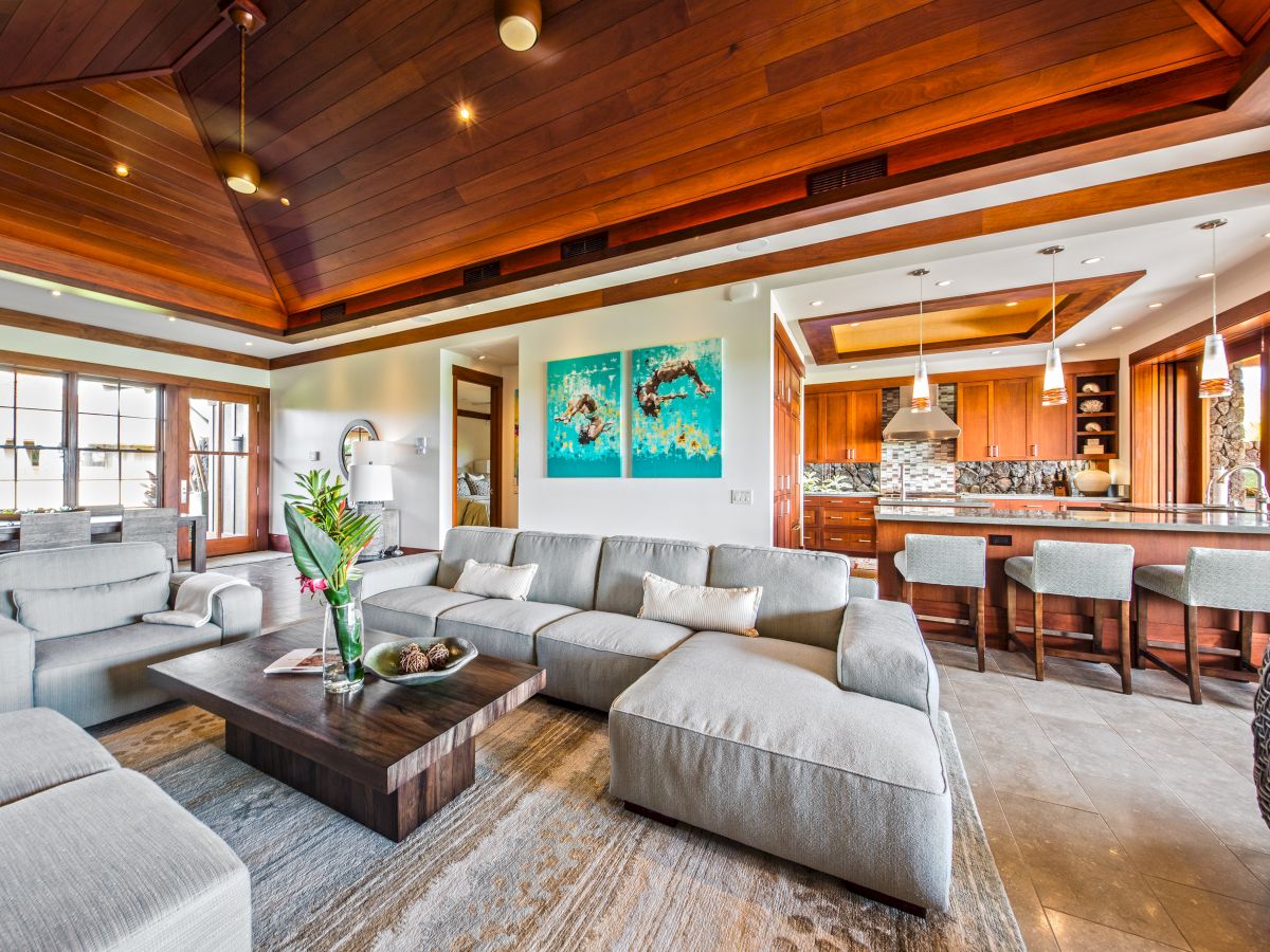Open-plan living room with gray sectional sofa, wood ceiling, pendant lights, and a modern kitchen island with stools.