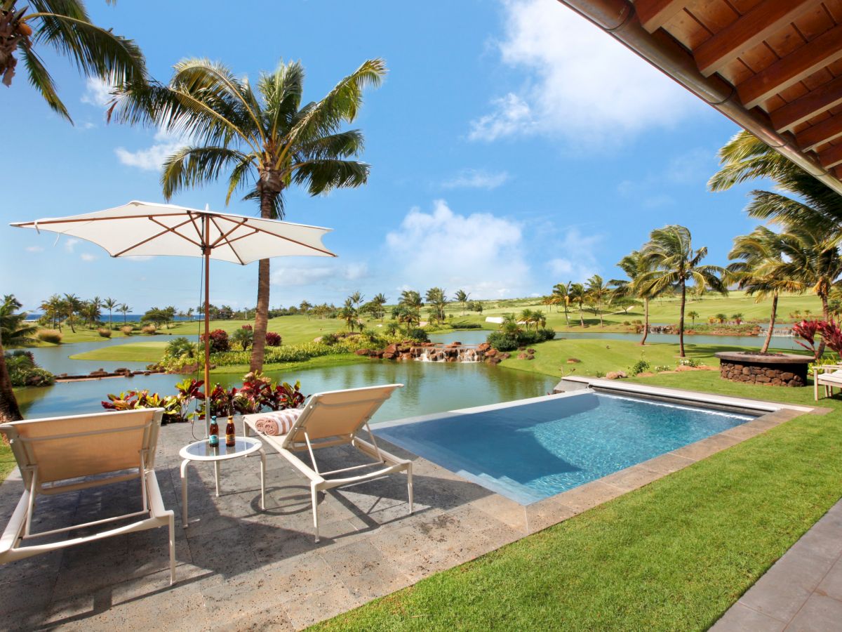 A sunny tropical resort scene with a pool, lounge chairs, an umbrella, palm trees, and a lake view with green lawn beyond.