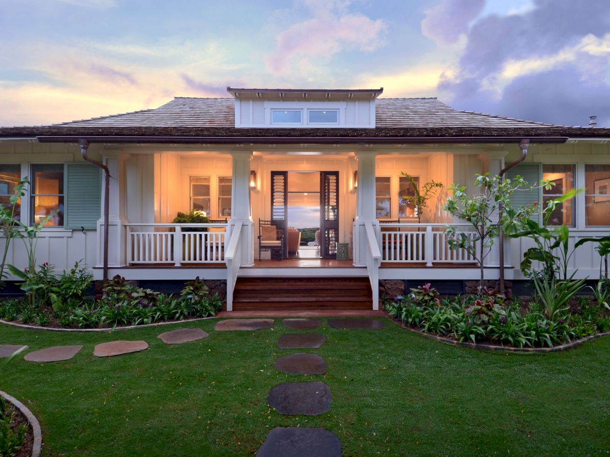 A modern white home exterior with a covered porch, railings, landscaped garden, and a sunset sky with colorful clouds.