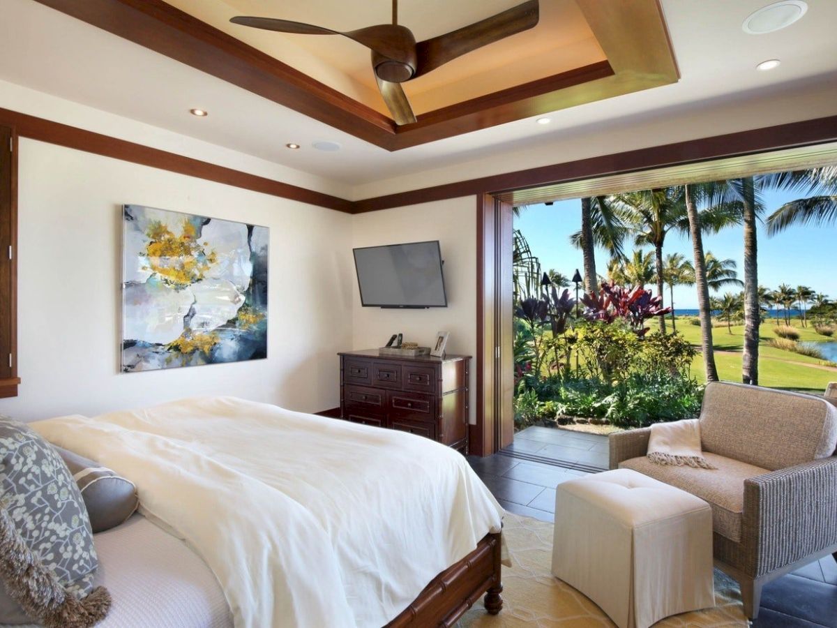 A sunny tropical bedroom with a ceiling fan, bed, TV, dresser, and open balcony view over palm trees and a lake.