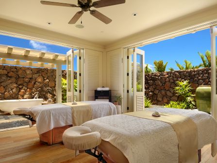A bright patio-style bedroom with a large bed, ceiling fan, open shutters, and a stone garden wall with tropical plants outside.