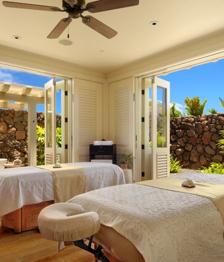 A bright patio-style bedroom with a large bed, ceiling fan, open shutters, and a stone garden wall with tropical plants outside.