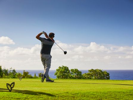 A golfer swings on a sunny course with the ocean and sky in the background.
