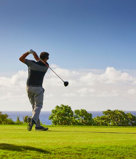 A golfer swings on a sunny course with the ocean and sky in the background.
