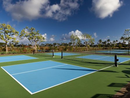 A sunny outdoor tennis court with a net, blue and green playing surface, and scattered trees under a dramatic blue sky with clouds.