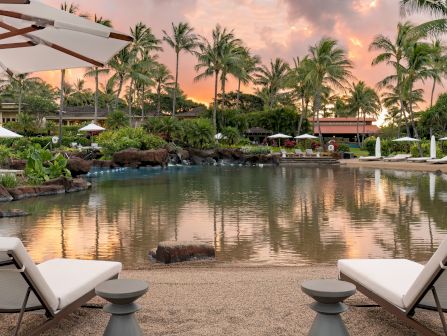 An elegant resort pool at sunset, ringed by palm trees and garden umbrellas, with loungers beside the calm water.