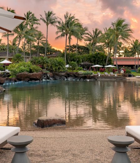 An elegant resort pool at sunset, ringed by palm trees and garden umbrellas, with loungers beside the calm water.