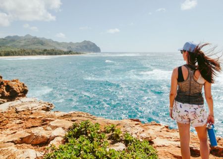 A woman in a cap stands by a rocky coast, looking over the turquoise ocean as the wind blows her hair.