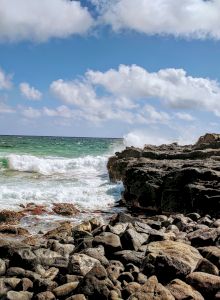 A windy coastline with churning green-blue waves crashing against dark rocky rocks under a sky full of fluffy clouds.