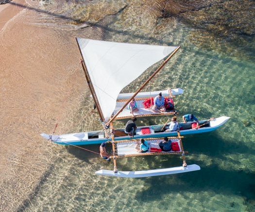 Aerial view of a catamaran sailboat with passengers near a sandy beach in clear blue-green water.